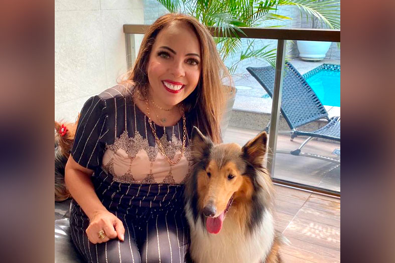 Edwiges Dias, mulher de pele branca com cabelos tingidos de ruivo. Ela está sorridente posando para foto ao lado de um cão da raça Collie.