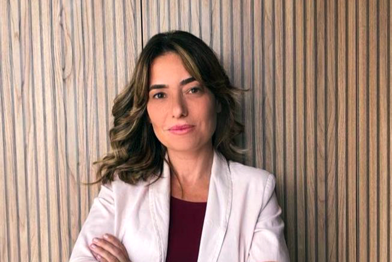 mulher de pele branca e cabelos castanhos claros posa para foto com os braços cruzados. Ela veste uma camisa vinho e um casado branco.