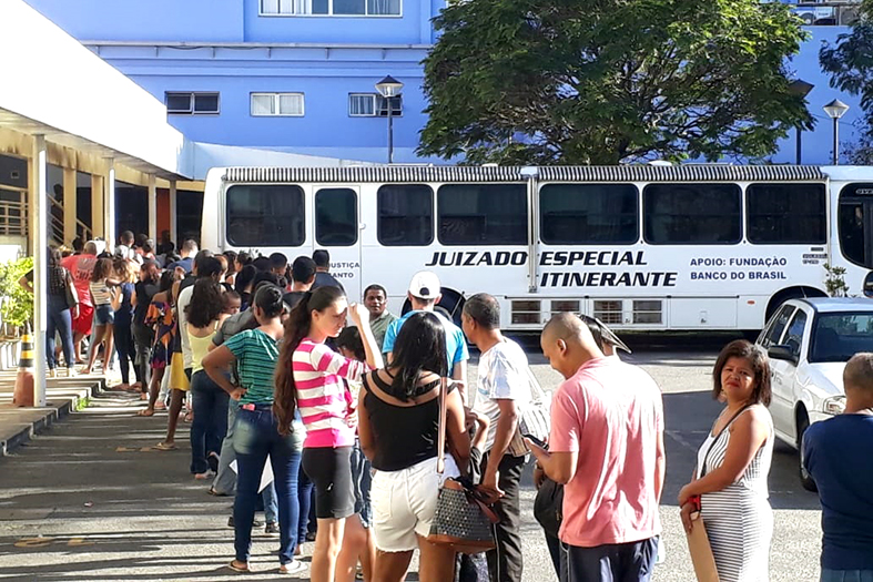 Fula de pessoas em direção a um ônibus branco com os seguintes dizeres em sua lateral; juizado especial itinerante.