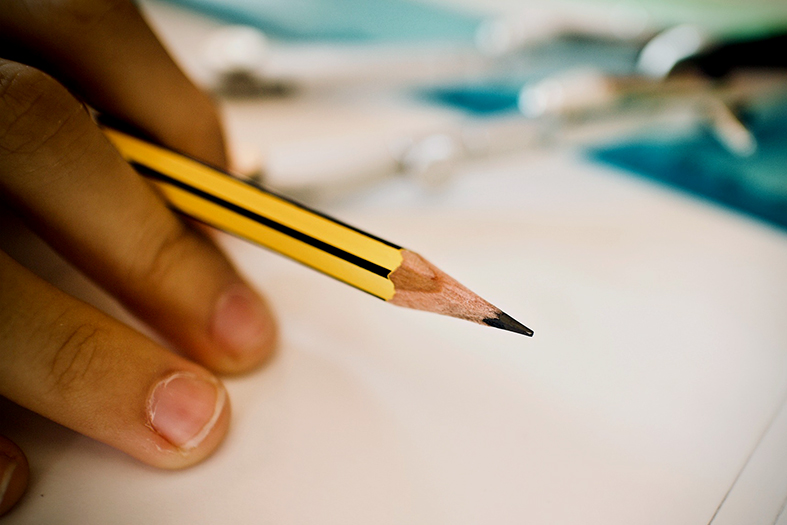 Detalhes de dedos segurando um lápis amarelo sobre um papel em branco.