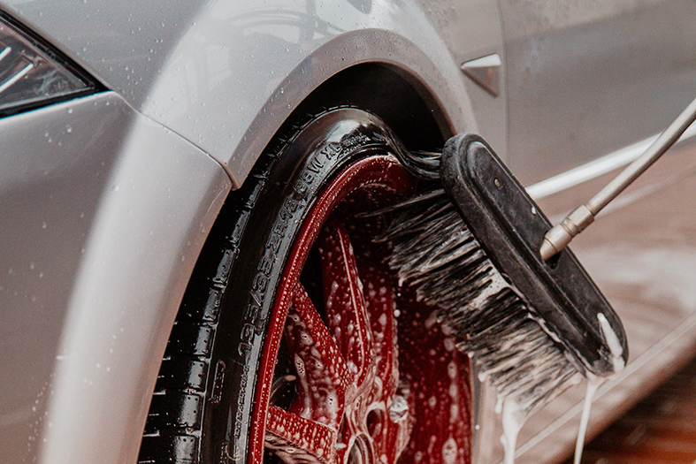 Detalhe de roda de carro sendo lavada com vassoura cheia de sabão.