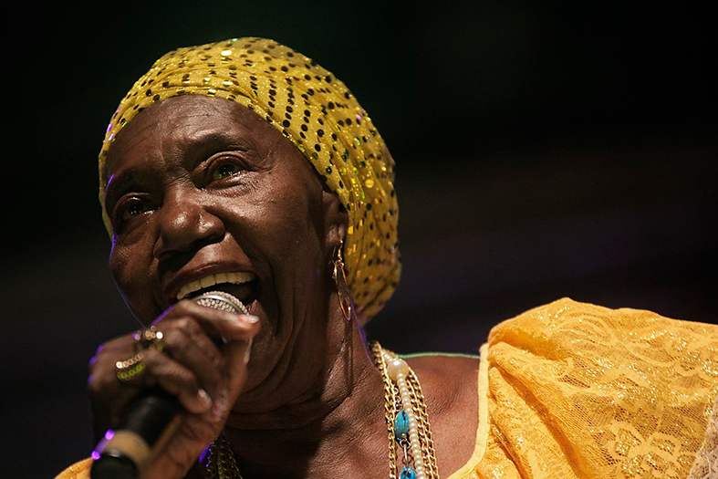 Lia de Itamaracá, mulher de pele negra, com vestido e turbante na cor amarela.