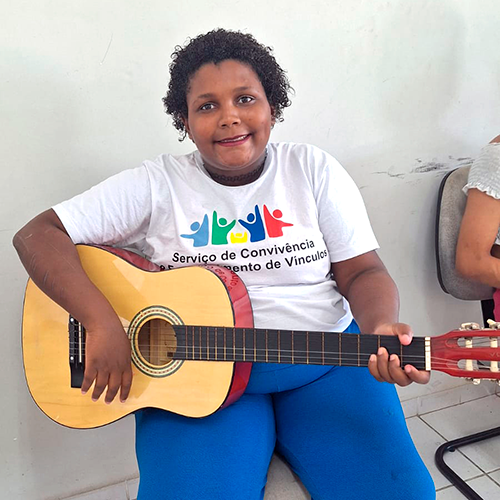 Maria Vitória, adolescente negra de 14 anos. Ela veste calça e camiseta e está segurando um violão.