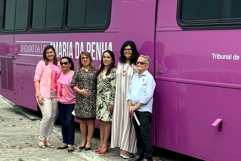 Cinco mulheres e um homem posam para foto em frente ao um ônibus do tipo turismo na cor rosa