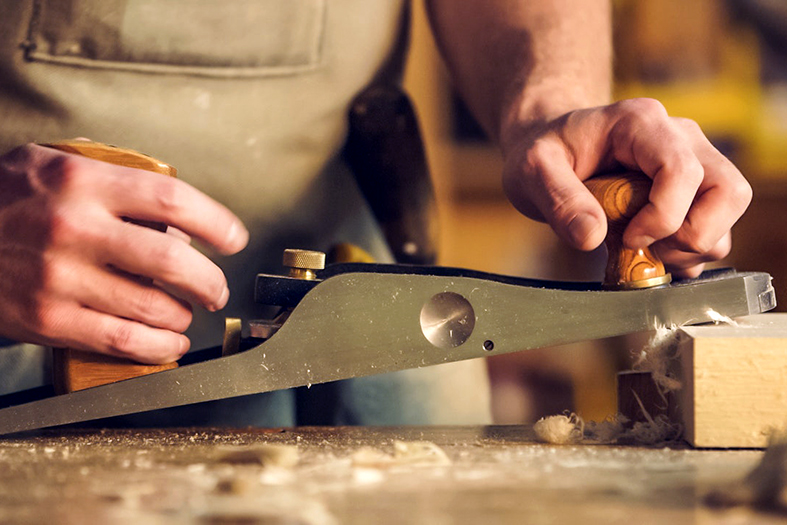 detalhe de um marceneiro desbastando uma peça de madeira com um instrumento manual.