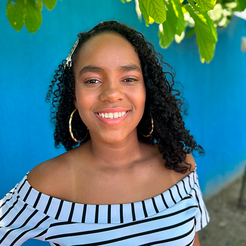 Maria Vitória, adolescente negra de cabelos anelados na cor preta. Ela veste uma blusa listrada e sorri para a câmera.