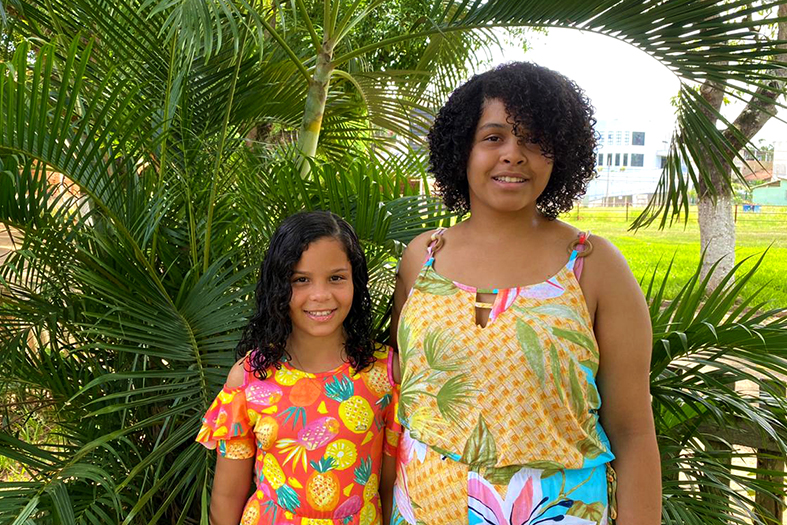 duas irmãs uma mais velha e mais alta do que a outra. a menor usa um vestido laranja com flores coloridas estampadas. a mais velha usa um vestido amarelo com estampas coloridas diversas.