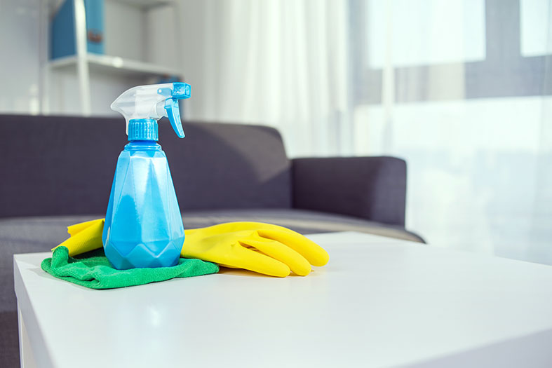 Em uma sala decorada com poucos móveis, um borrifador com líquido azulado, luvas amarelas e pano verde repousam sobre mesa de centro na cor branca.