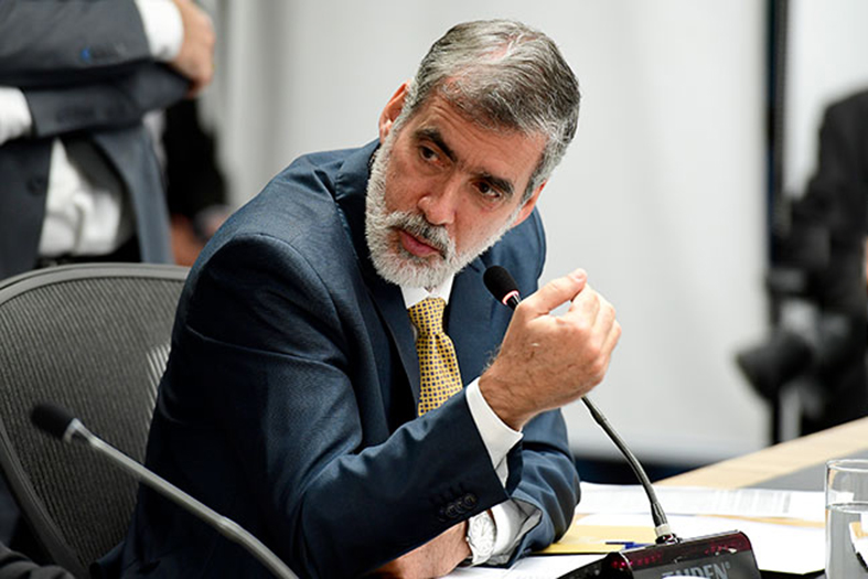 homem de pele branca, cabelos e barba grisalhos, vestindo terno azul marinho fala ao microfone.
