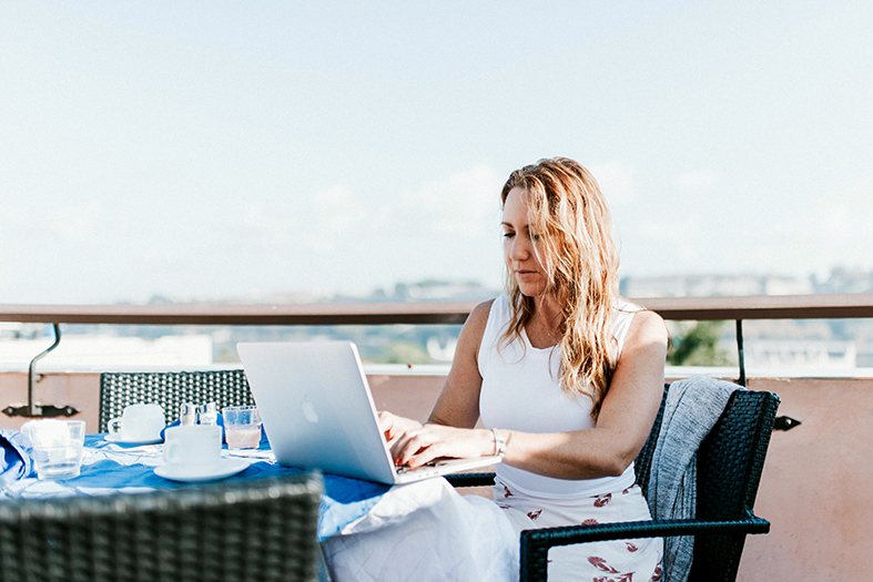 Mulher de pele branca e cabelos loiros digitando em um laptop apoiado em uma mesa posta de café da manhã.