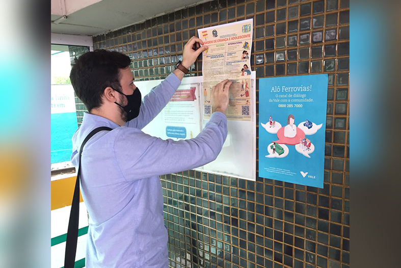 Homem de estatura alta, com pele branca e cabelos na cor preta, coloca um cartaz na parede.