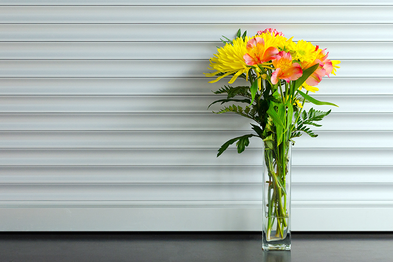 Persianas de cor branca ao fundo e vaso de virdro longo com flores de variadas cores em primeiro plano