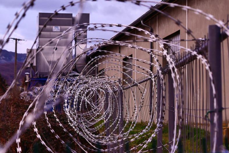 Arame de concertina enrolado protegendo o topo de um muro de prisão. ao fundo uma guarita.