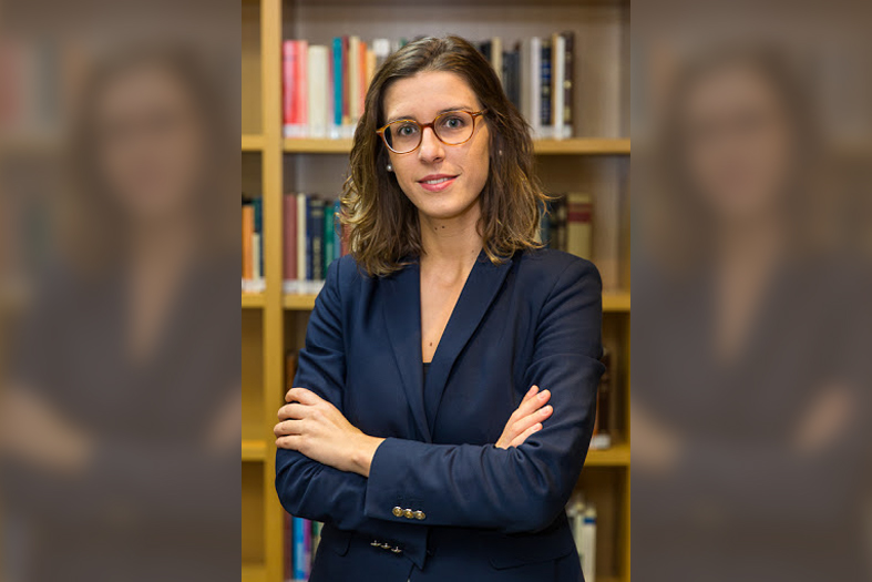 professora sofia temer posa para foto trajando um terno do tipo tailleur na cor azul escuro. Ao fundo uma estante de livros.