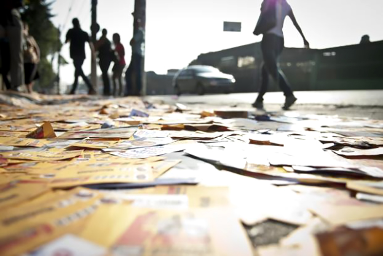 Vários "santinhos" ou propagandas de candidatos às eleições do ponto de vista de baixo para cima. Ao fundo silhuetas de pessoas andando.