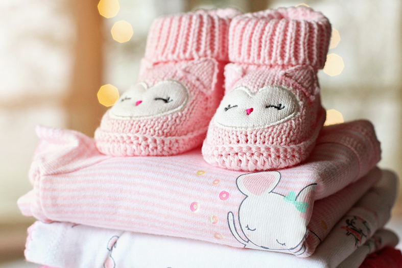 Sapatinhos de tecido para bebês na cor rosa repousam sobre mudas de roupas em tons de rosa.