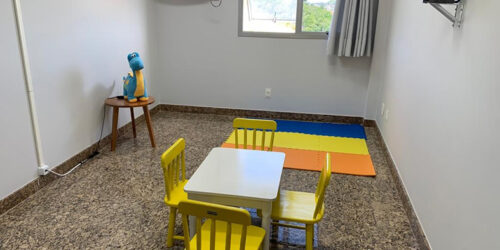 sala pequena com decoração simples contando com uma mesinha infantil na cor amarela e em uma mesa de cabeceira um dinossauro de pelúcia na cor azul. no alto da parede esquerda uma câmera de vigilância