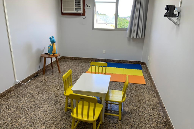 sala pequena com decoração simples contando com uma mesinha infantil na cor amarela e em uma mesa de cabeceira um dinossauro de pelúcia na cor azul. no alto da parede esquerda uma câmera de vigilância