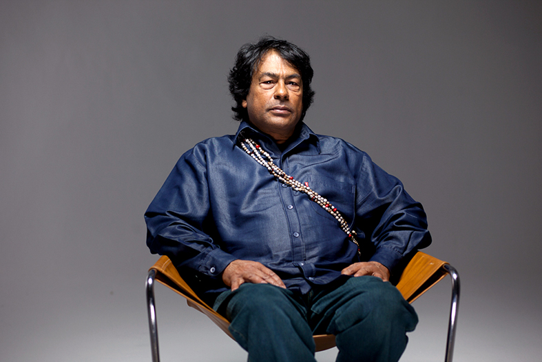 Ailton Krenak, indígena, cabelos pretos, está sentado em uma cadeira e veste blusa e calças cor jeans.