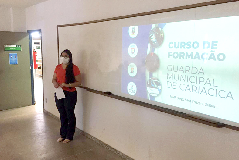mulher de pele branca, estatura mediada, de cabelos pretos e longos. ela está em pé em frente a um quadro branco onde é projetado uma apresentação. o título da apresentação, projetado, no quado branco é: curso de formação - guarda municipal de cariacica