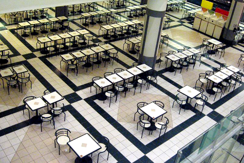 Salão vasto com inúmeras mesas com quatro cadeiras cada sugerindo uma praça de alimentação típica de shoppings.