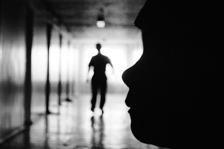 fotografia em preto-e-branco de um corredor onde se vê duas silhuetas. Um rosto infantil de perfil em primeiro plano. Uma forma humana em segundo plano se movimentando pelo corredor.