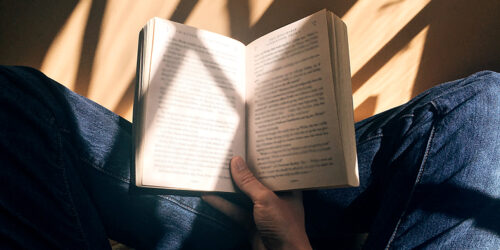 Detalhe de uma pessoa jovem do sexo masculino e de pele branca lendo um livro apoiado em seu colo. Ele está de pernas cruzadas e usa calças jeans.