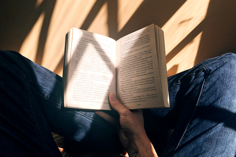 Detalhe de uma pessoa jovem do sexo masculino e de pele branca lendo um livro apoiado em seu colo. Ele está de pernas cruzadas e usa calças jeans.