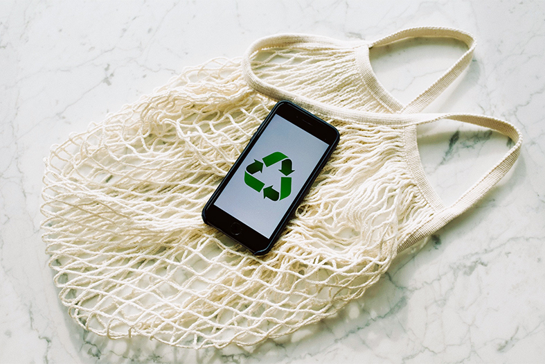 Um celular com a tela exibindo o símbolo da reciclagem repousa sobre uma bolsa de compras de tecido trançado.