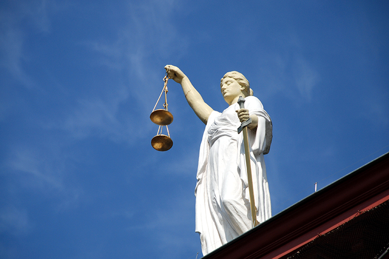 Estátua da deusa themis vista de um ângulo de baixo para cima. a estátua é branca, ela segura uma balança dourada. a estátua está posta em cima de uma estrutura alta.