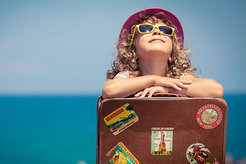 Menina de óculos escuros se apoia sobre uma mala de viagem.