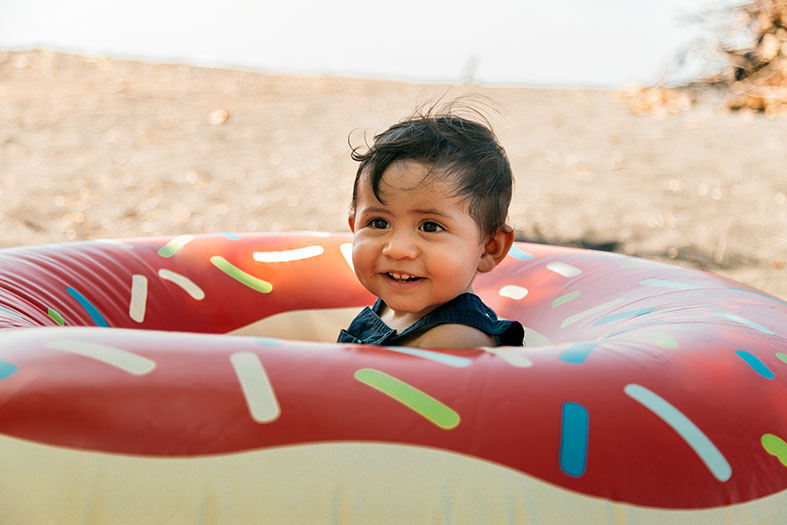 Em uma praia, criança de colo brinca dentro de uma boia decorada como uma rosquinha.