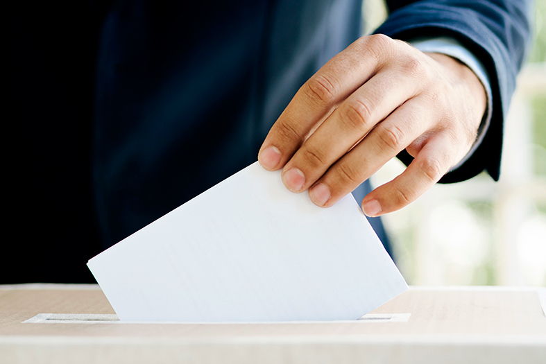 mão masculina coloca papel branco em uma urna de papelão. A pessoa veste terno azul marinho.